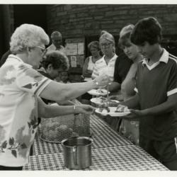 Fish Boil: Volunteer, Marge Clink, serving food behind table to visitors outside Visitor Center