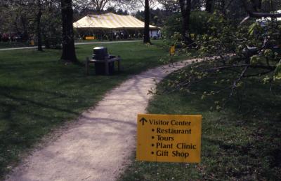 Arbor Week directional sign to Visitor Center next to walking path with tent in background