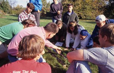 Pat Kelsey and students seated outside on ground reviewing soil trench for Wheaton College soil class