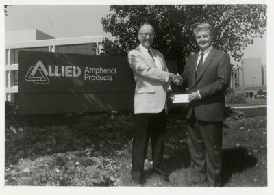 Dr. Marion Hall receiving check and shaking hands with man in front of Allied Amphenol Products