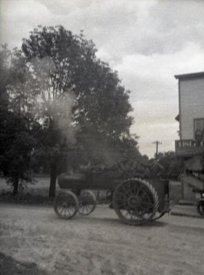 Man driving Lisle Farms threshing outfit in front of Riedy's drug store in Lisle (out of focus)