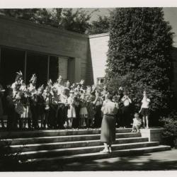 May Watts leading group of children wearing feather headbands in musical performance on steps outside of Thornhill