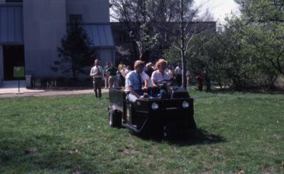 Dr. Tom Green and Karla Patterson in cart leading line of people across lawn near Research building for tree planting at Arborfest