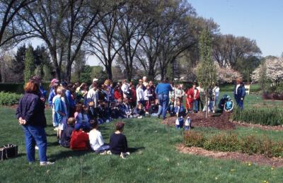 Deb Seymour assisting crowd, mainly children, with shovels at Arbor Day tree planting near Hedge Garden