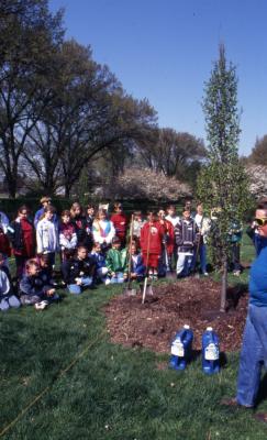 Deb Seymour near Arbor Day planted tree with crowd, mainly children, gathered around near Hedge Garden