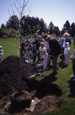 Crowd watching little girl with shovel and man with hat planting tree on Arbor Day