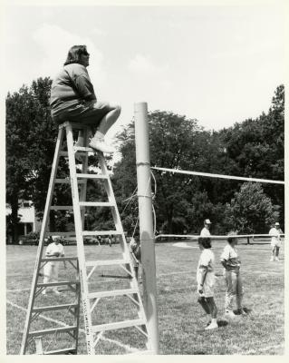 Deb Seymour, referee, seated on ladder looking over employee summer picnic volleyball game near the Research Building