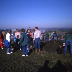 Crowd on top of berm with mulch pile at right and camera stand at left during Earth Day celebration and berm planting