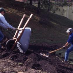 Two grounds staff dumping soil from wheel barrel and raking soil near lake